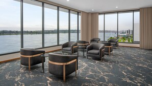 A hotel room with large windows overlooking a body of water and a bridge, featuring several grey upholstered chairs and a patterned carpet.