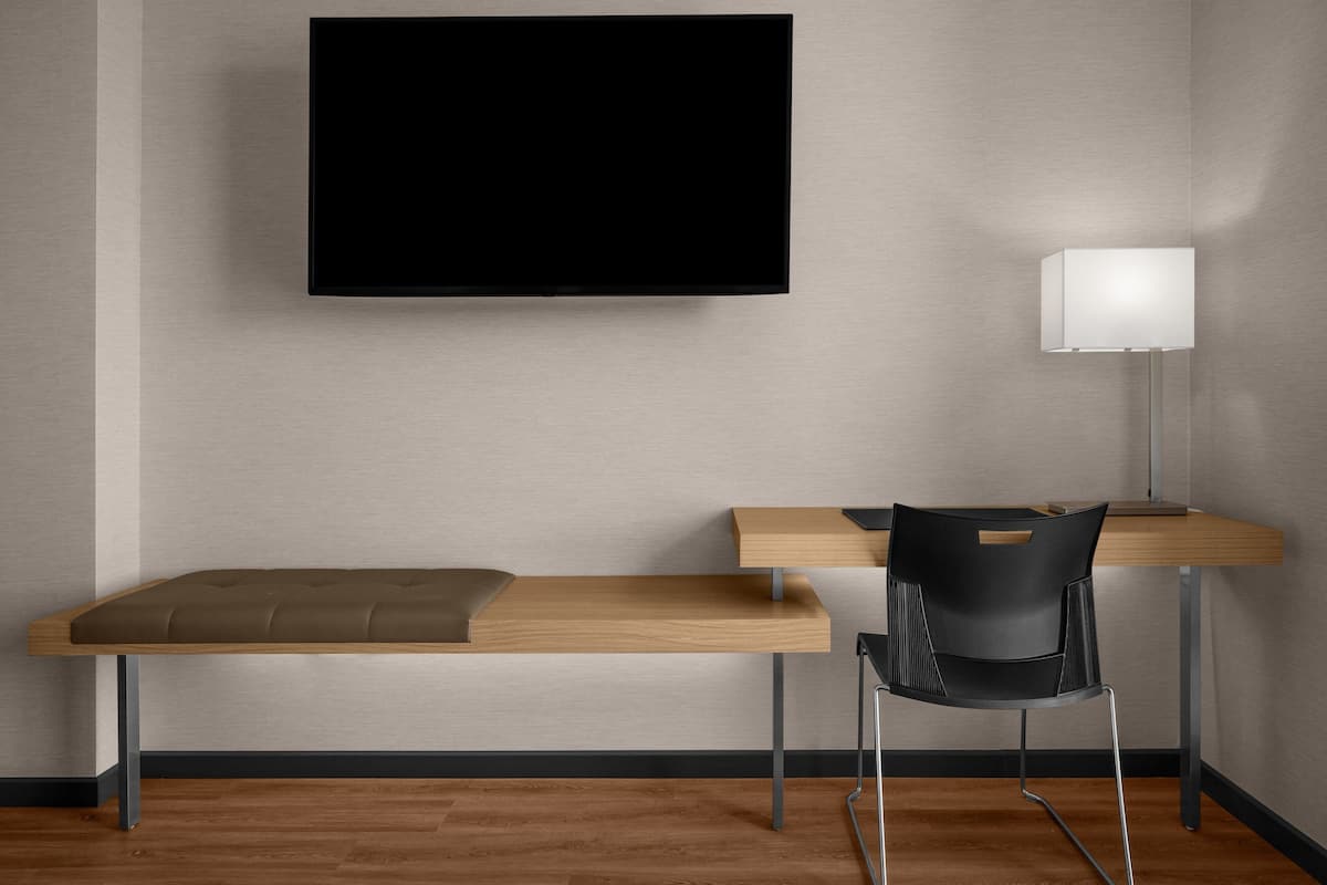 A modern room with a flat-screen TV mounted on the wall, a wooden desk with a bench, a black chair, and a floor lamp.