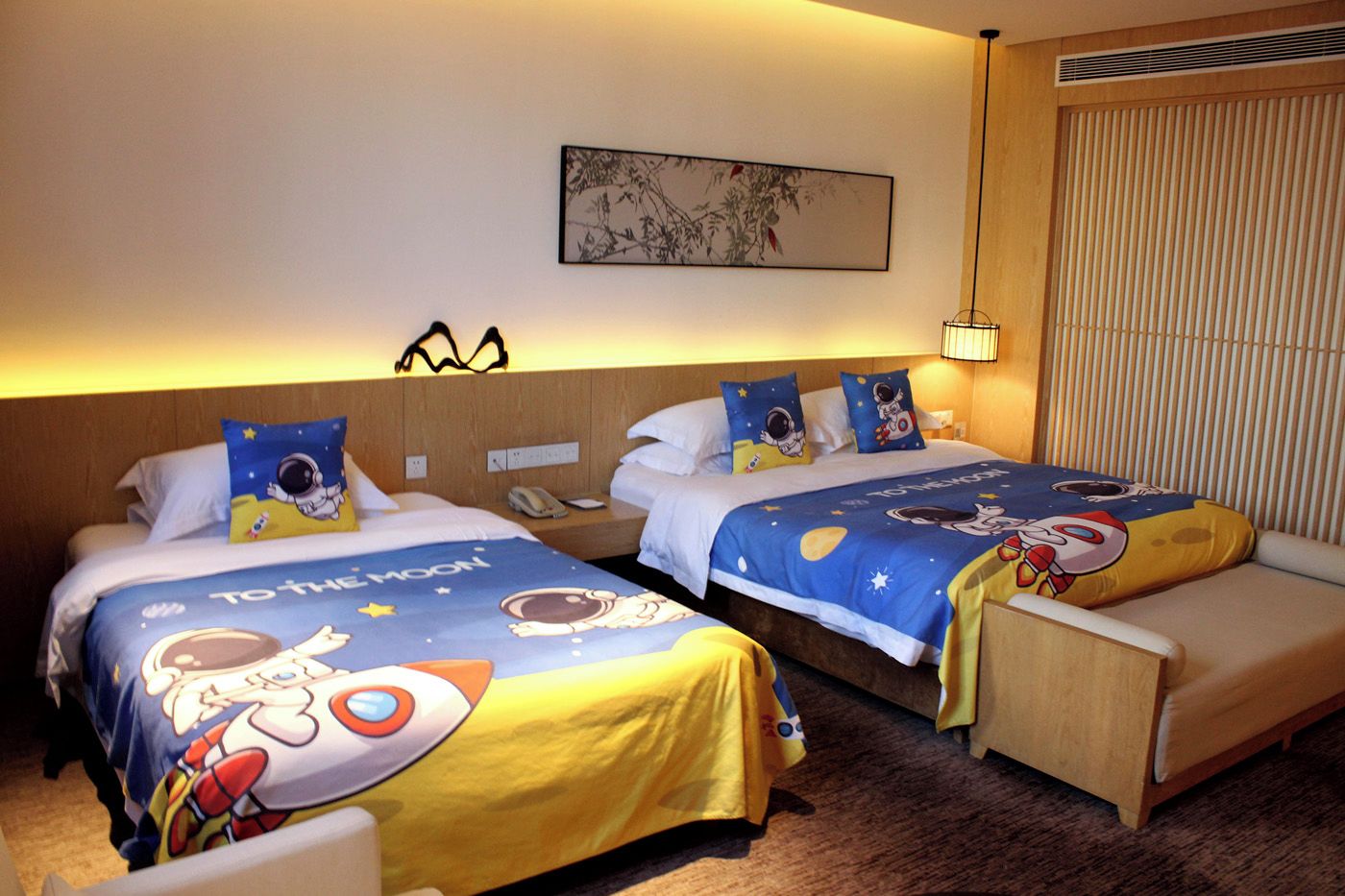 A hotel room with two beds, each with a space-themed bedspread, a bedside table with a lamp, and a wall-mounted artwork.