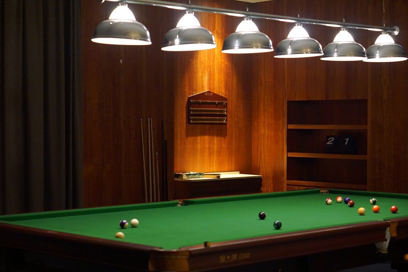 Una mesa de billar con bolas, iluminación colgante y una pared de madera con estante y tablero de números.