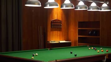 Una mesa de billar con bolas, iluminación colgante y una pared de madera con estante y tablero de números.