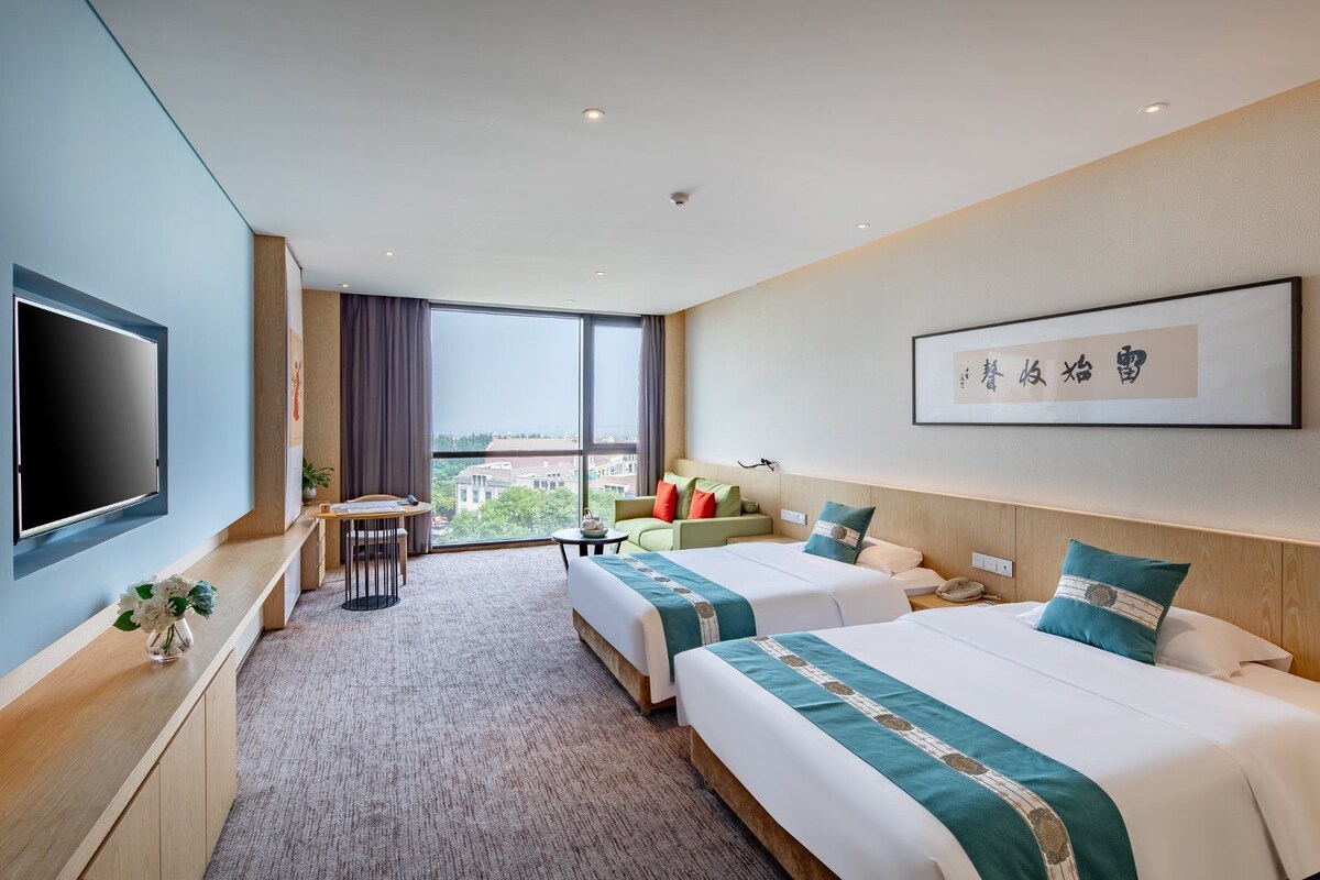 A hotel room with two beds, a TV, a desk, and a large window offering a city view.