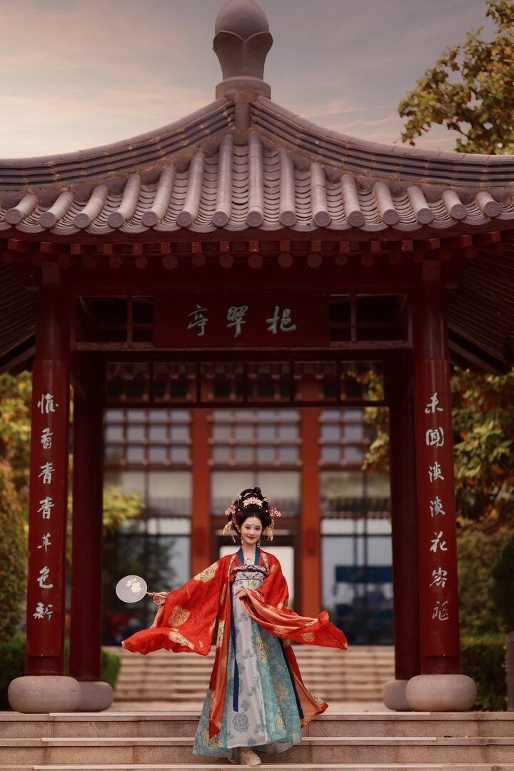 A traditional Chinese pavilion with a tiled roof and red pillars, featuring calligraphy and a person in traditional attire.