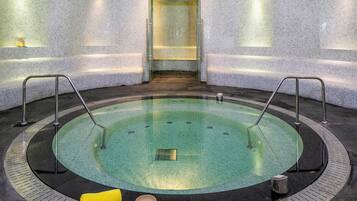 Een rond bubbelbad met glazen wanden, omgeven door een betegelde vloer en muren, met een bankje en een kaars op de vloer.