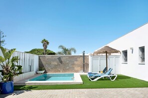 Une piscine avec des chaises longues, un parasol en chaume et un mur en pierre.