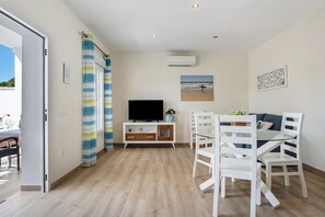 Ein Zimmer mit Fernseher, Esstisch mit Stühlen, einem Fenster mit Vorhängen und einer Wandklimaanlage.