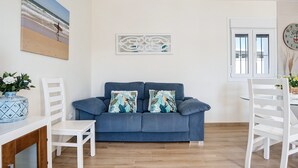 Ein Wohnzimmer mit einem blauen Sofa, weißen Stühlen, einer Holzkommode, einem Strandbild und einer Wanddekoration.