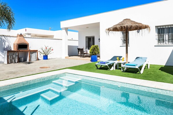 Ein Swimmingpool mit Liegestühlen, einem Grill und einem Strohdachschirm.