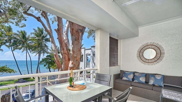 A balcony with a table, chairs, and a sofa, overlooking a beach with palm trees and the sea. - Absolute Beach Front 1 Bedroom Apartment Palm Cove (Cairns)
