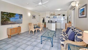 A living room with a glass coffee table, wicker furniture, a kitchen area with a fridge, microwave, and sink, and a ceiling fan. - Absolute Beach Front 1 Bedroom Apartment Palm Cove (Cairns)