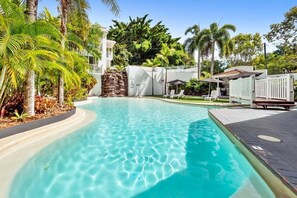 A swimming pool with clear water, surrounded by palm trees and a white building. - Alassio 302-2 | Beachfront Studio (Cairns)