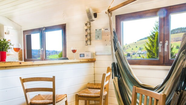Une pièce accueillante avec un hamac, des chaises en bois, une petite table sur laquelle repose un verre de vin, et une vue sur un paysage verdoyant par de grandes fenêtres.