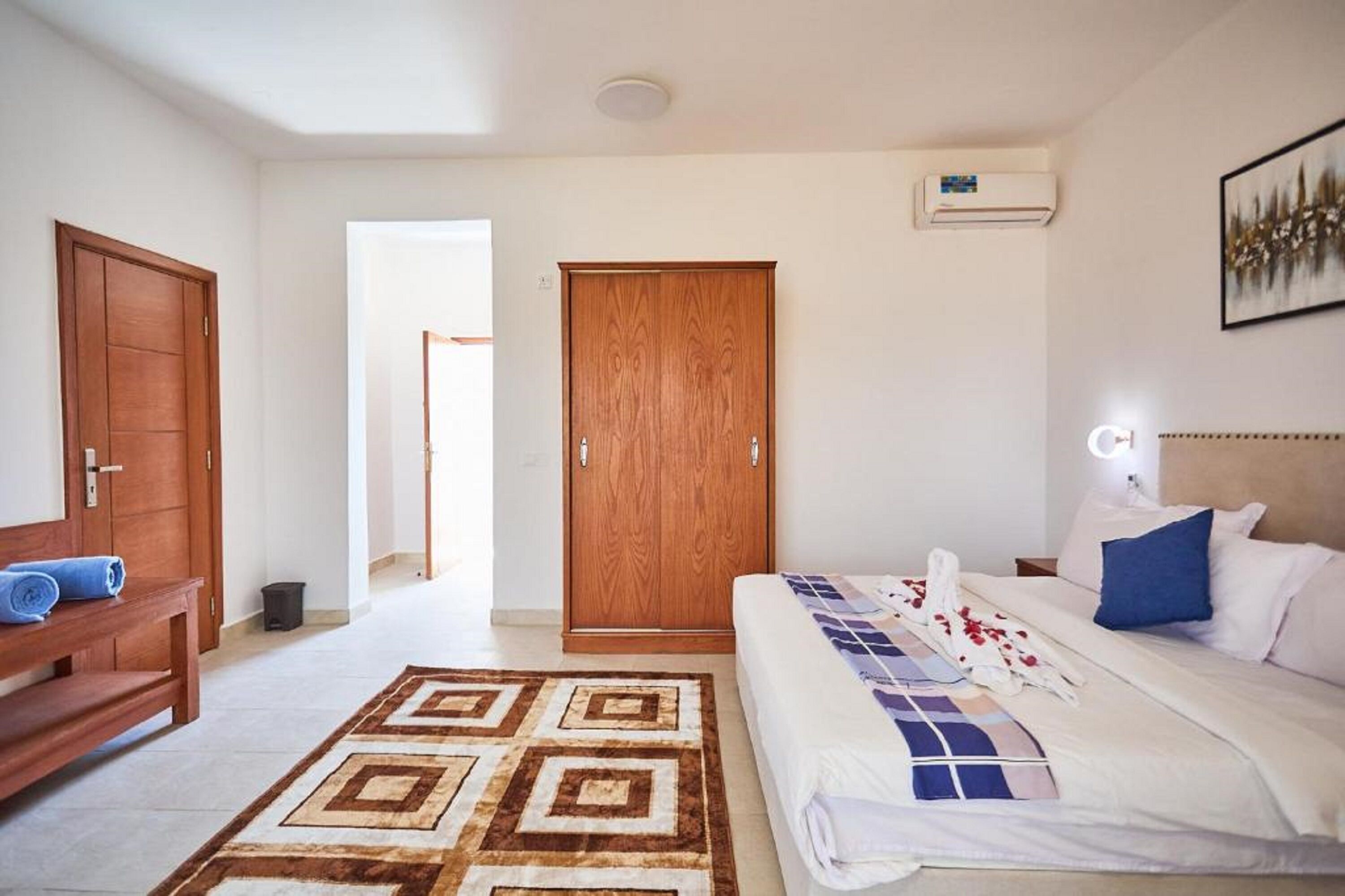 Hotellihuone, jossa on sänky, puinen vaatekaappi, penkki, kuviollinen matto ja taulu seinällä.