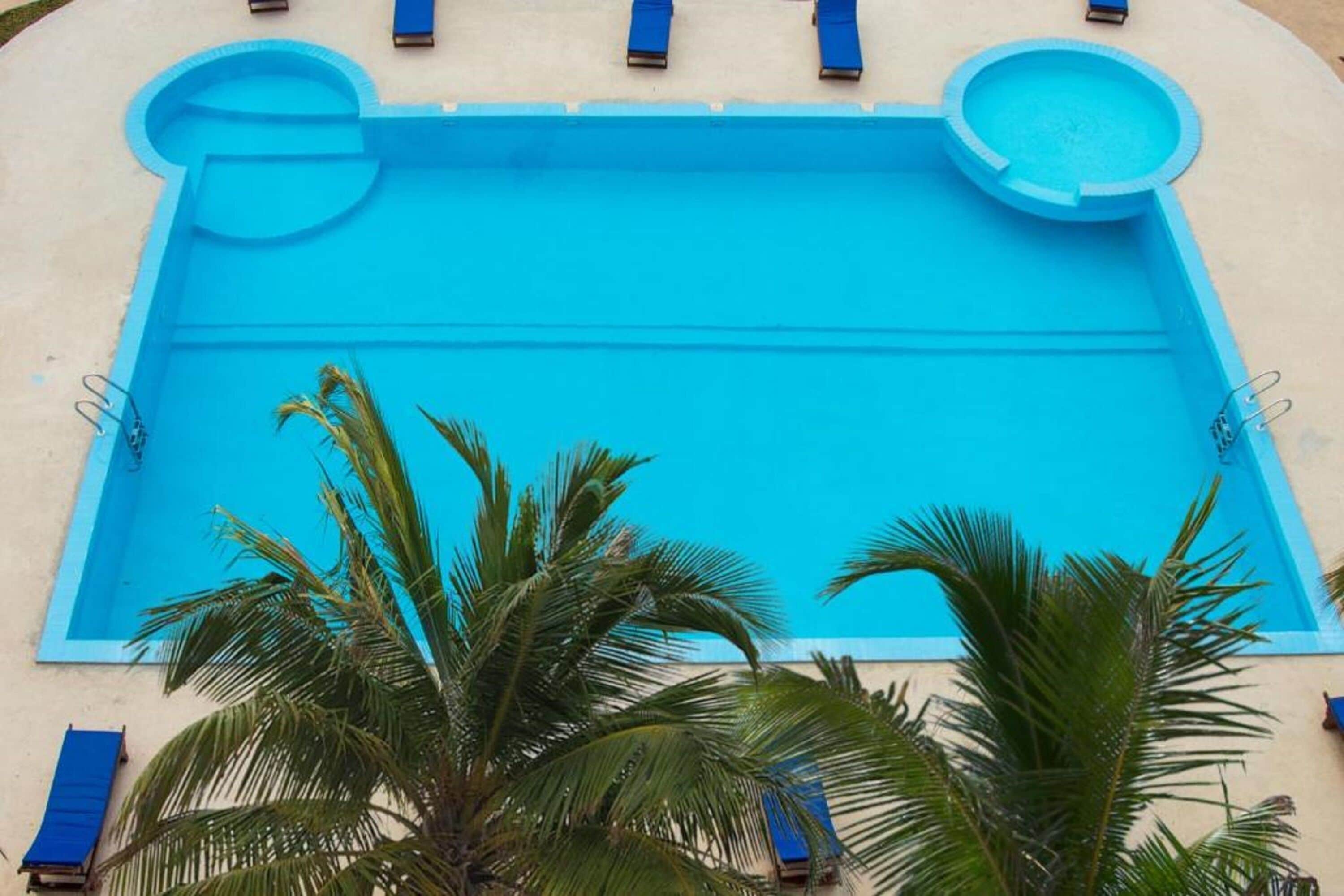 Una piscina rectangular con revestimiento azul, rodeada por un muro de color claro. En cada extremo de la piscina hay dos jacuzzis circulares, y se pueden ver algunas tumbonas. Palmeras en primer plano.