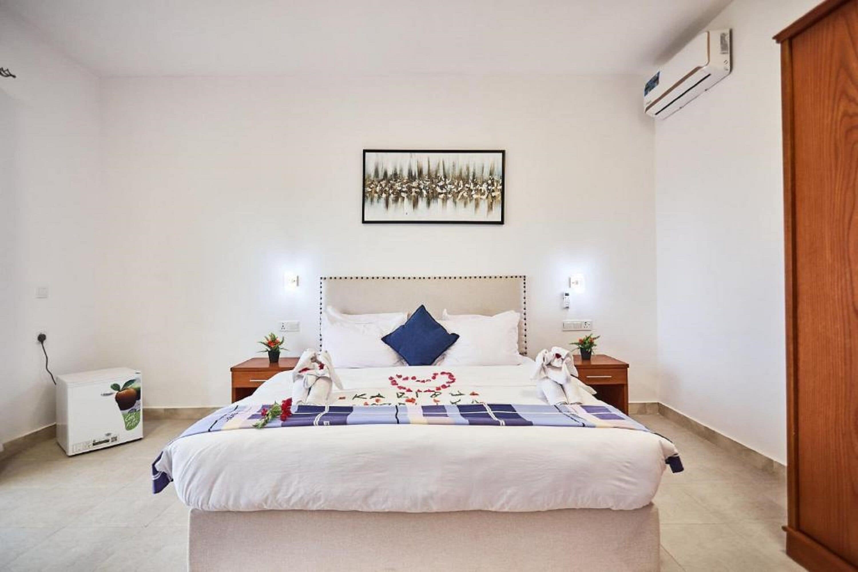 Una cama bien hecha con un adorno con forma de corazón, un cuadro en la pared y una pequeña mesa con una planta.