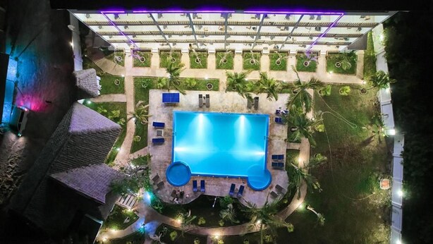 An aerial view of a hotel with a central swimming pool, surrounded by palm trees and outdoor seating areas.