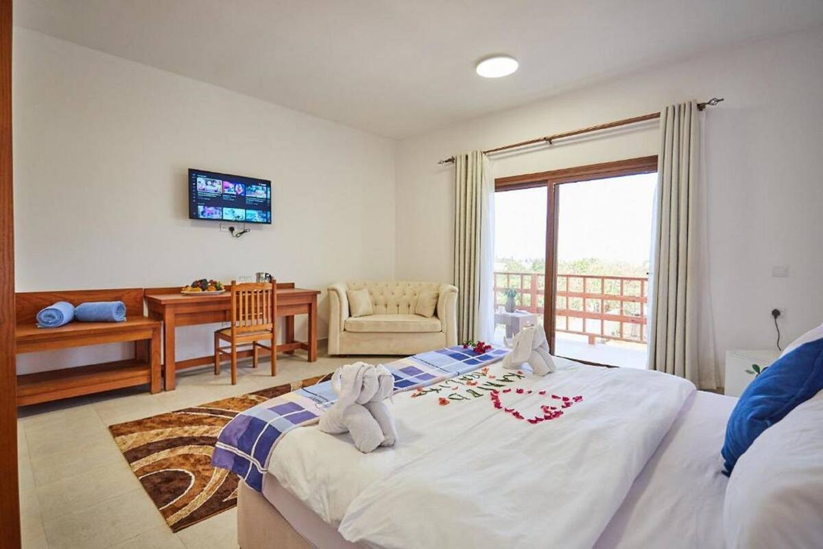 Habitación de hotel con cama, escritorio, silla, televisor y balcón con cortinas.