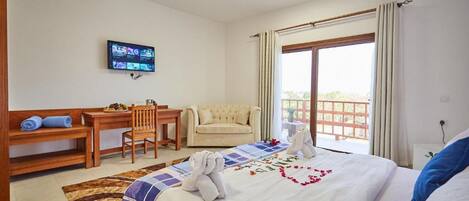 Habitación de hotel con cama, escritorio, silla, televisor y balcón con cortinas.