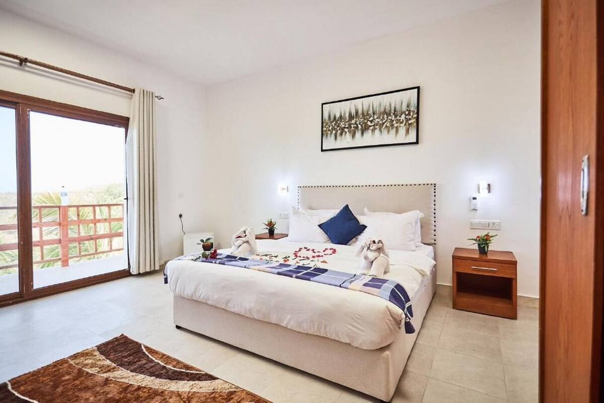 Un dormitorio con una cama grande, una mesita de noche de madera, un cuadro enmarcado en la pared y vistas a través de la puerta de cristal.