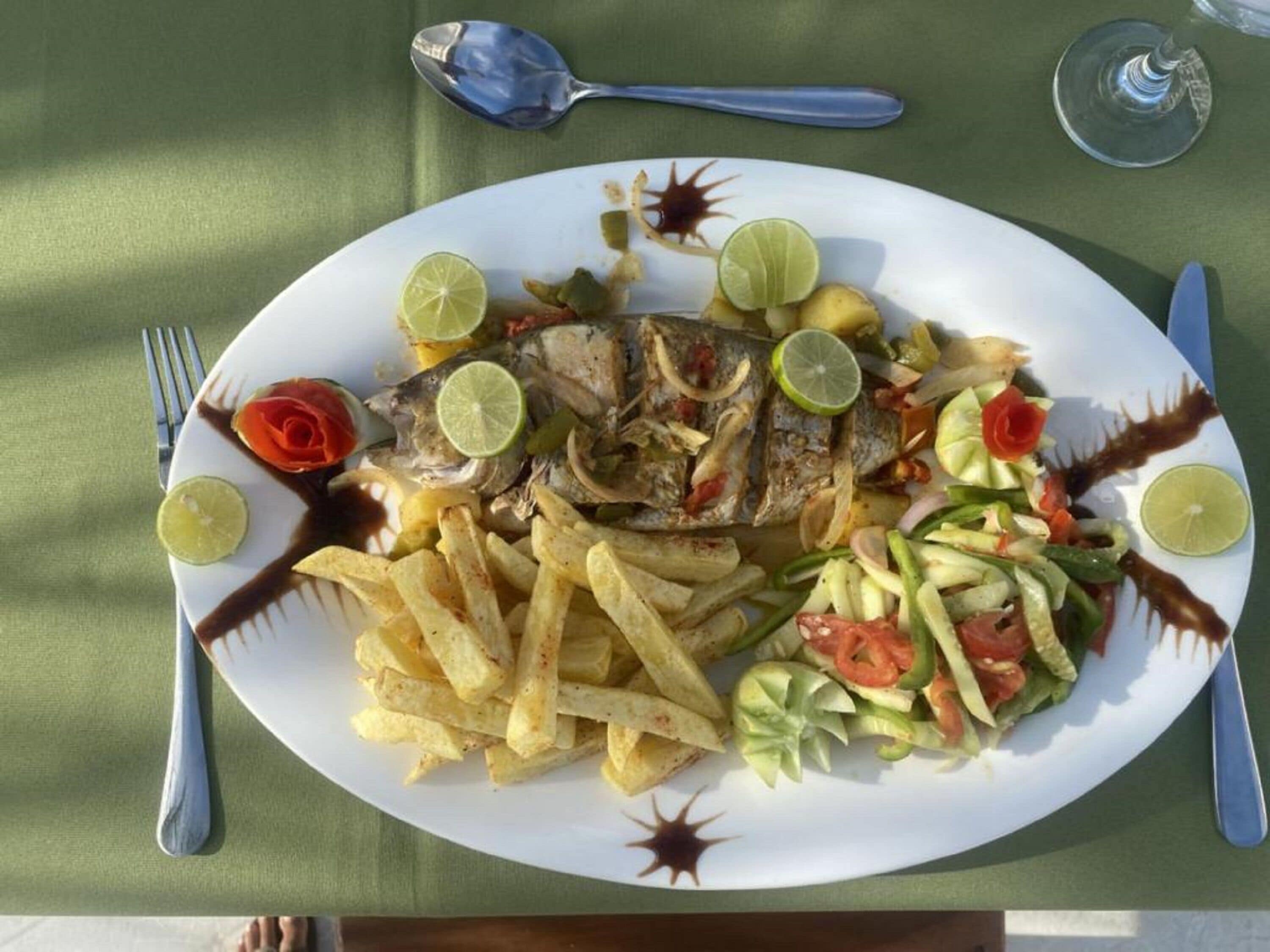 A plate of food with fish, fries, and a salad, accompanied by a spoon and a glass.