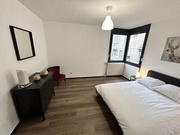 A bedroom with a bed, a chair, a dresser, a radiator, and a window with a view of buildings.