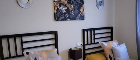 A hotel room with two beds, each with a headboard and a yellow bedspread. There is a wall-mounted artwork above the beds and a small table with a lamp between them.
