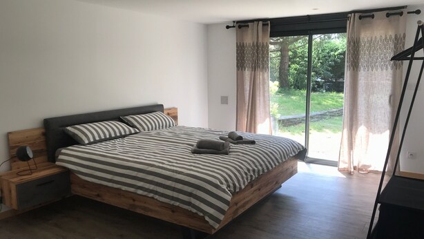 A bedroom with a wooden bed, striped bedding, a nightstand, and a view of greenery through a sliding glass door. - 4-bedroom family house with sauna, garden and terrace - near south beach (Lacanau)