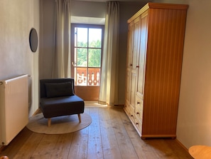 Et værelse med trægulv, en mørk sofa, en træskab og et vindue med udsigt til grøn bevoksning.