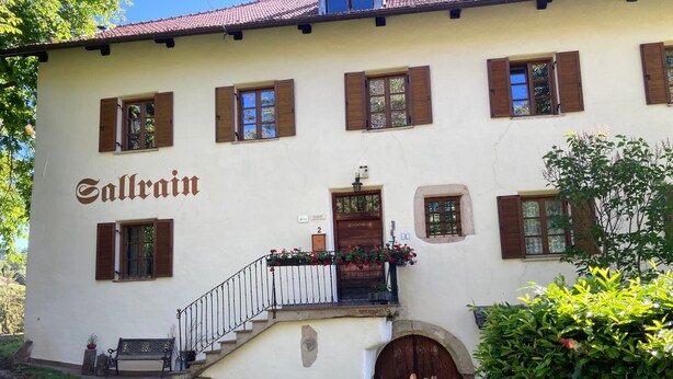 Ein Gebäude mit der Aufschrift 'Sallrain' an der Fassade, einem Holzzaun und einer Bank vor der Tür.