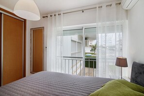 Un dormitorio con cama, una ventana con cortinas, una lámpara de noche y vistas al exterior.