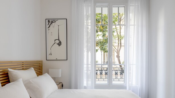 A hotel room with a bed, a nightstand, a lamp, a framed artwork, and a view of a balcony with greenery. - Almond Blossom, by Veeve (Paris)