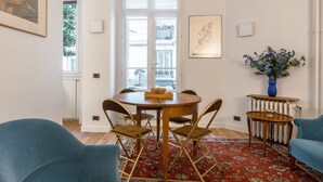 A dining area with a round table, four chairs, a red patterned rug, a blue vase with flowers, and a window with a view of buildings. - Almond Blossom, by Veeve (Paris)