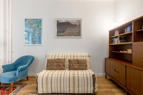 A room with a striped sofa, a blue armchair, a wooden bookshelf, and two framed pictures on the wall. - Almond Blossom, by Veeve (Paris)