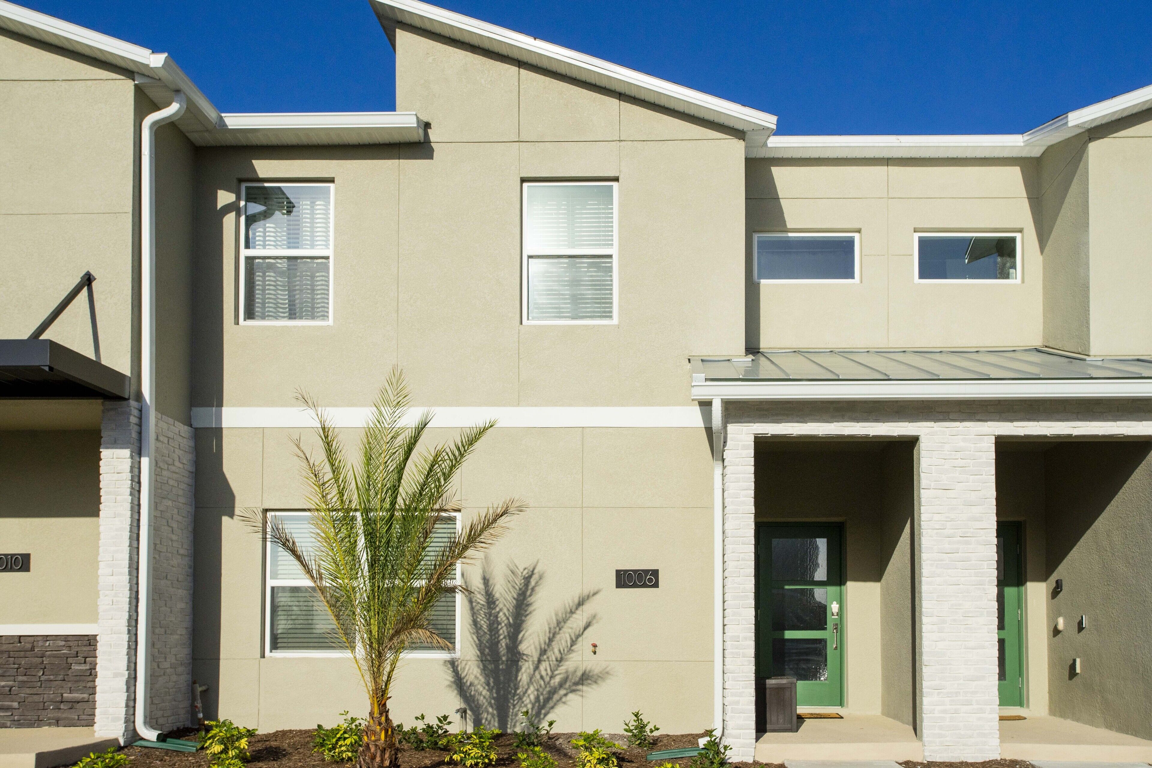 A modern apartment building with a flat facade, numbered 1006, featuring a green door and a small garden area with a palm tree.