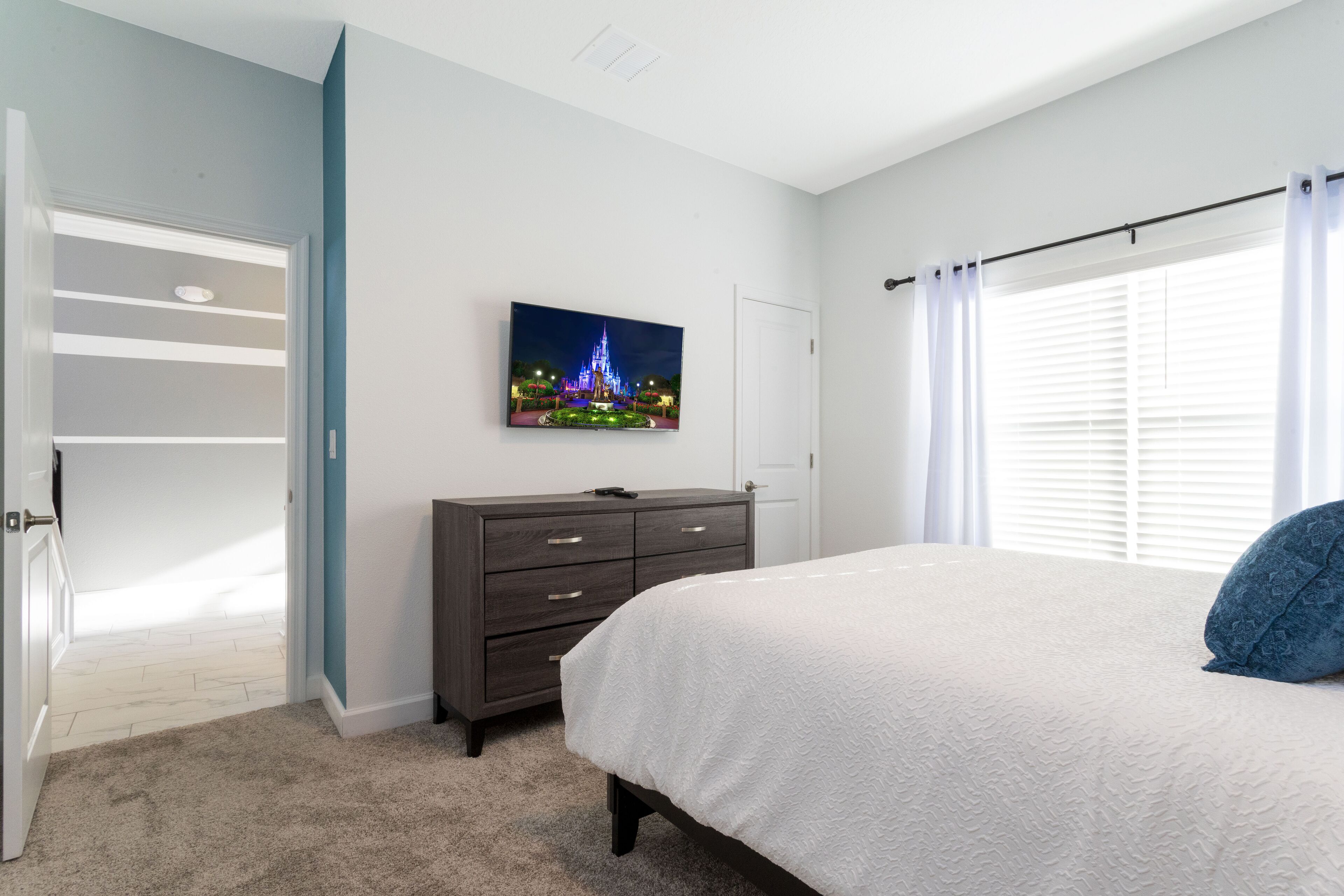 A bedroom with a bed, a dresser, a door, and a wall-mounted TV displaying a nighttime scene.