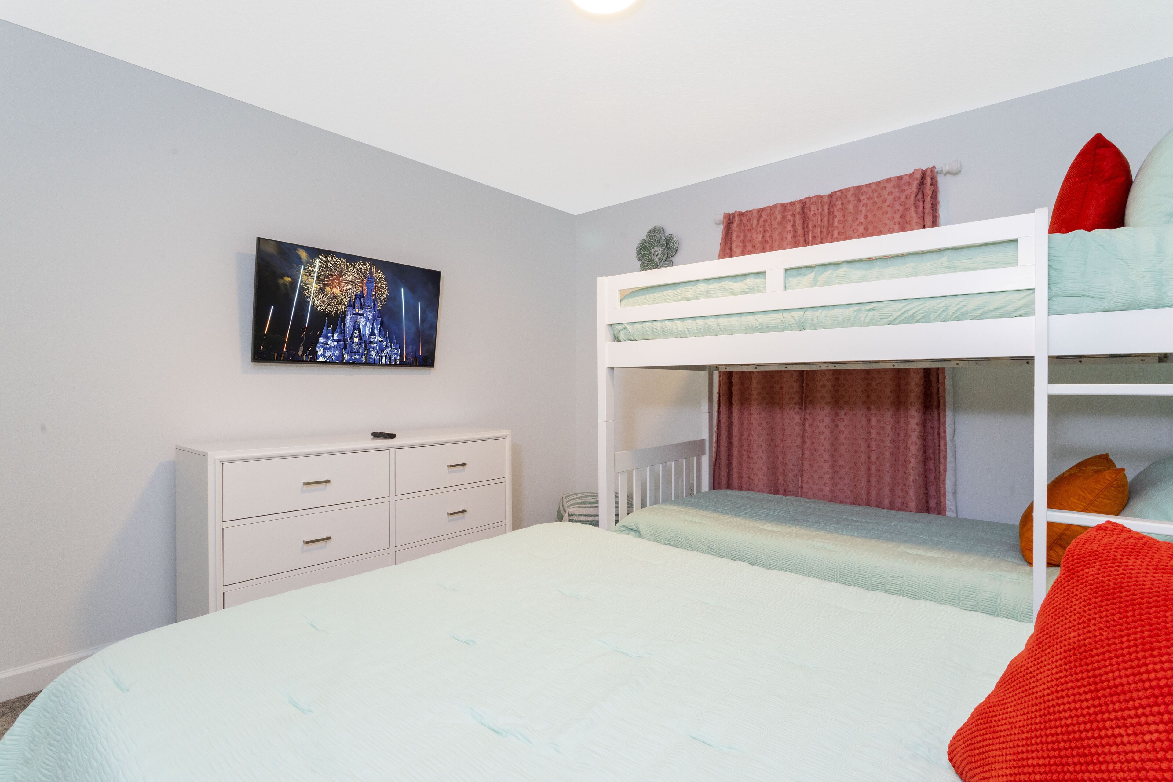 A bedroom with a bunk bed, a single bed, a white dresser, and a wall-mounted TV displaying fireworks.