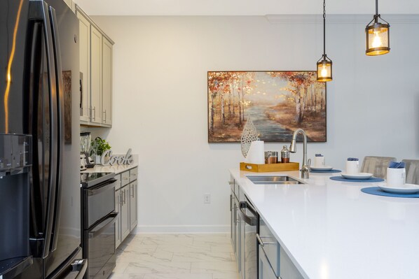 Una cocina moderna con un refrigerador de acero inoxidable, armarios blancos, una encimera blanca y un cuadro con una escena de bosque.