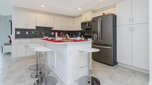 A modern kitchen with white cabinets, a stainless steel refrigerator, a microwave, and a central island with bar stools. - Gorgeous 3Bd 8 Guests @Storey Lake Resort Close to Disney 101 3191 (Kissimmee)