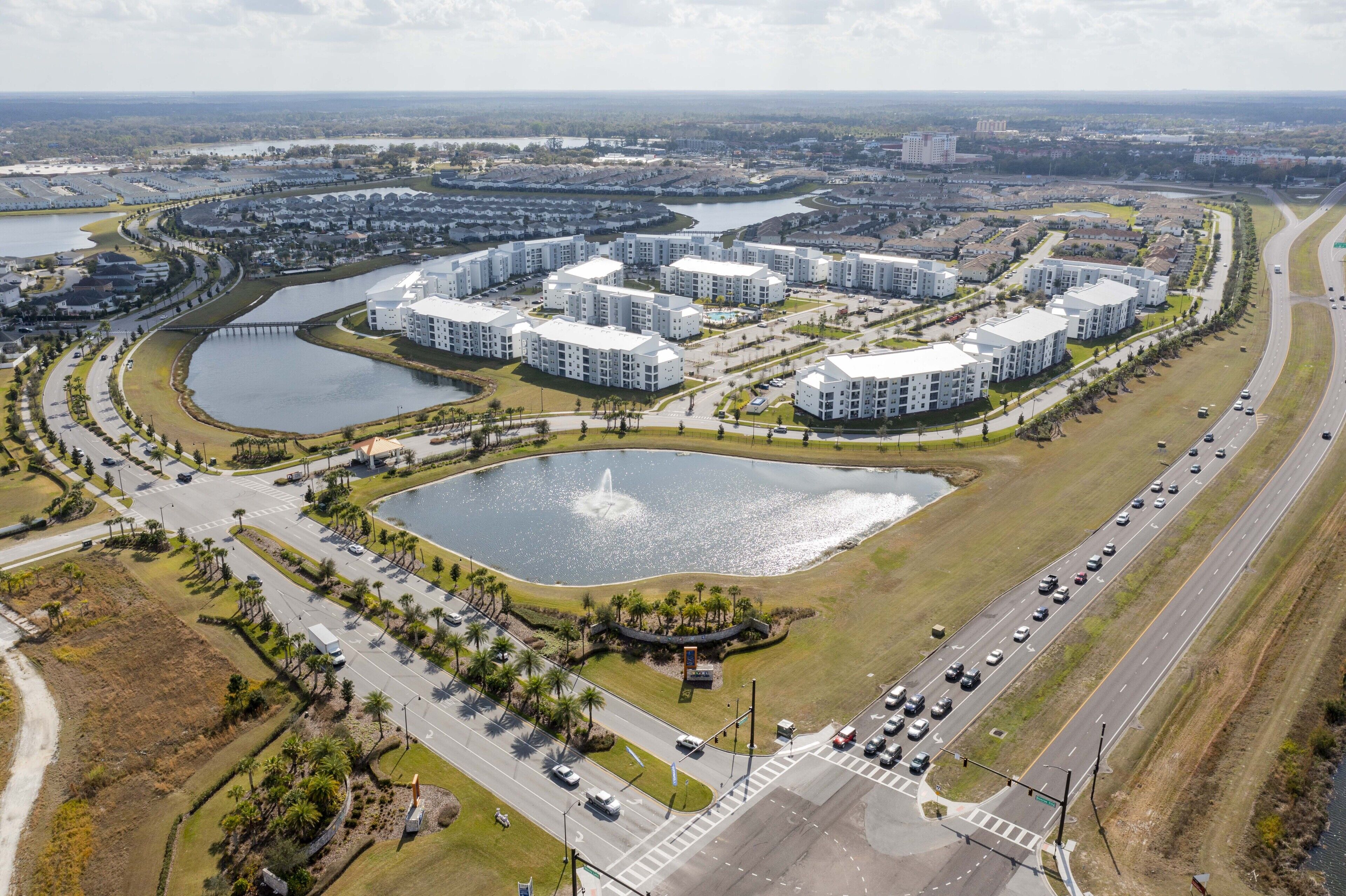 An aerial view of a residential area with modern apartment buildings, a large central water feature, and a surrounding road with vehicles.