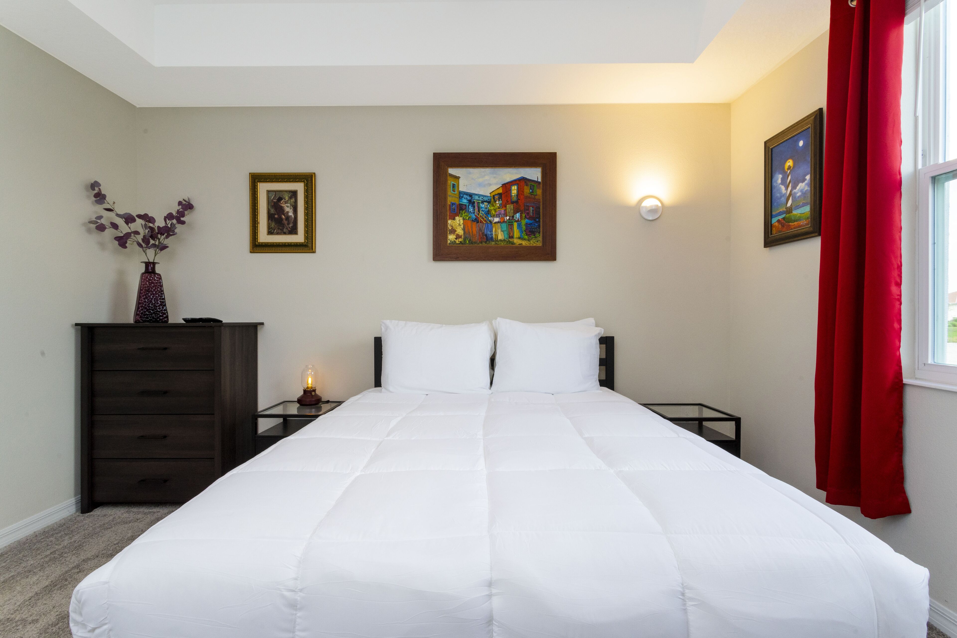 A neatly made bed with white bedding, a dresser with drawers, a nightstand with a lamp, and two framed artworks on the walls.