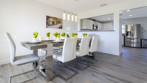 Una cocina moderna con electrodomésticos de acero inoxidable, una zona de comedor con sillas blancas y una mesa de madera, y un cuadro en la pared.