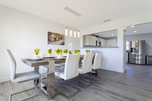 Eine moderne Küche mit Edelstahlgeräten, ein Essbereich mit weißen Stühlen und einem Holztisch sowie ein Gemälde an der Wand.