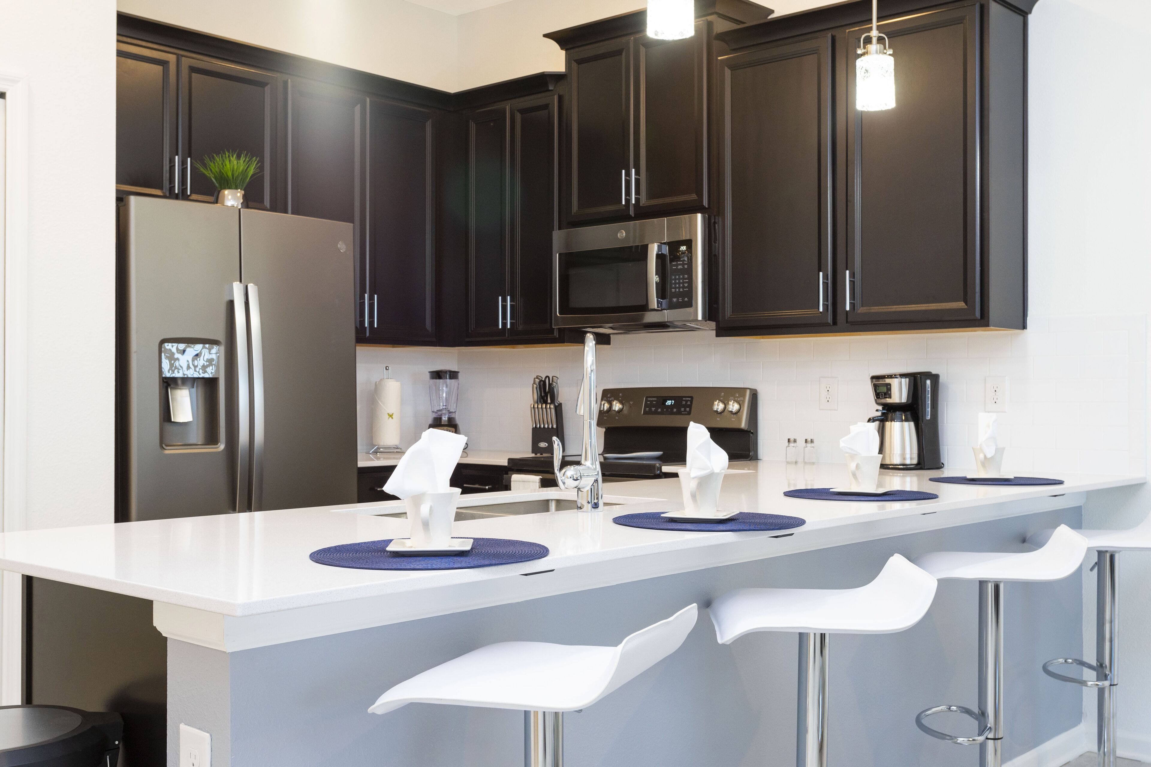 A modern kitchen with dark cabinets, a white countertop, stainless steel appliances, and bar stools.