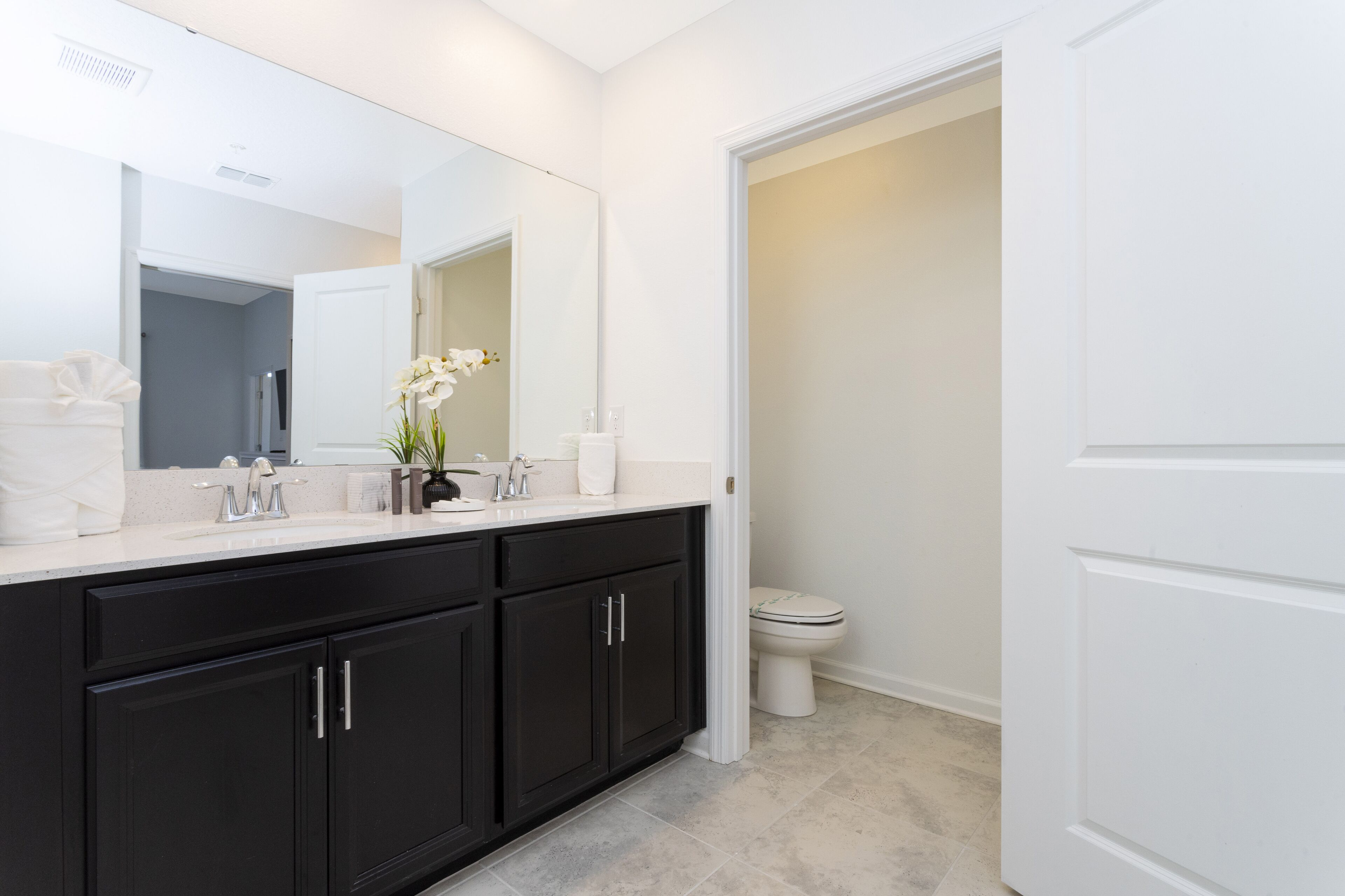 A modern bathroom with a large mirror, dark cabinets, a white toilet, and a sink with a vase of flowers.