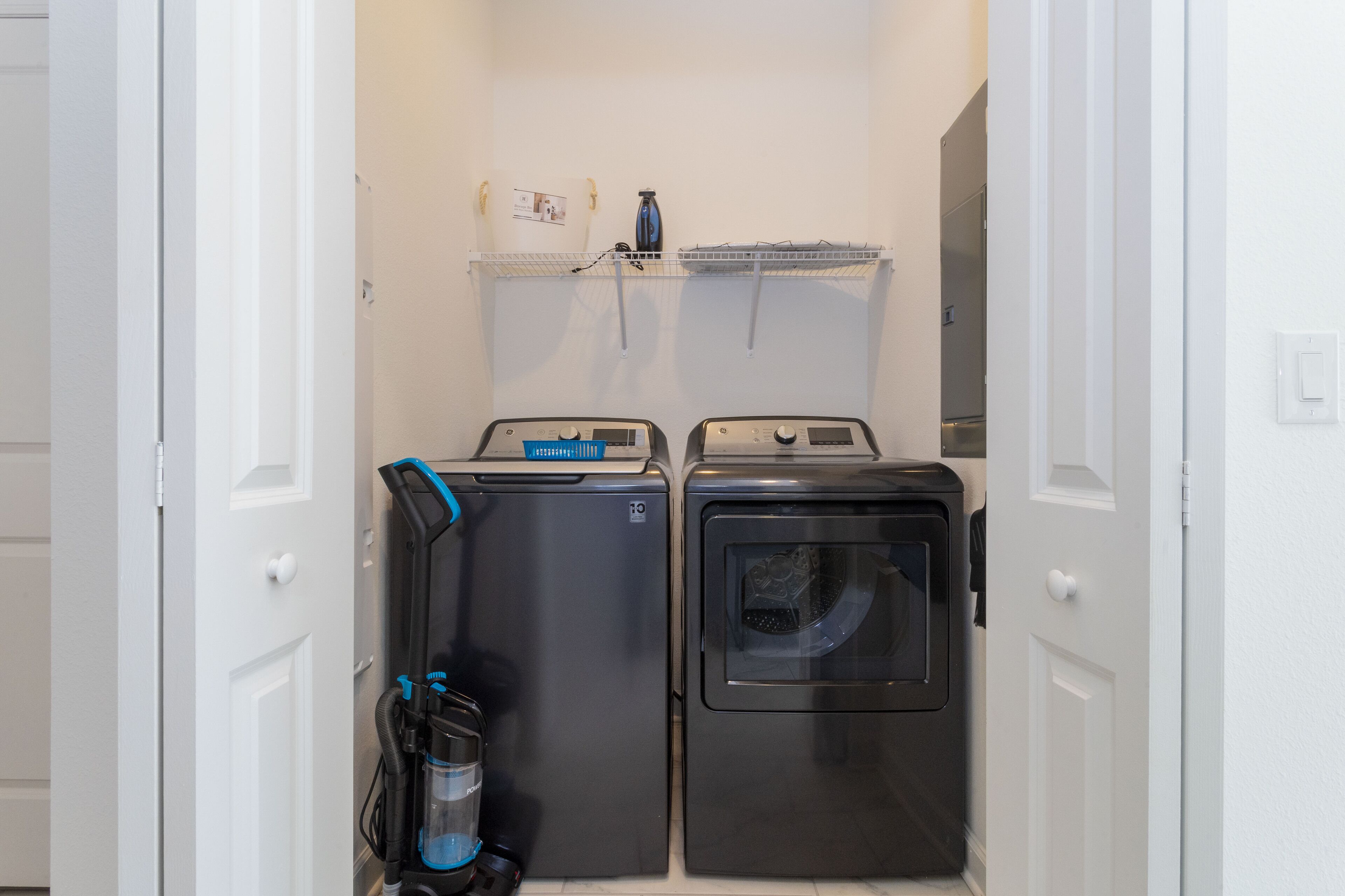 A laundry room with two washing machines and a dryer, a water heater, and a shelf with a water bottle and papers.