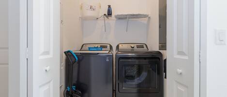 A laundry room with two washing machines and a dryer, a water heater, and a shelf with a water bottle and papers.