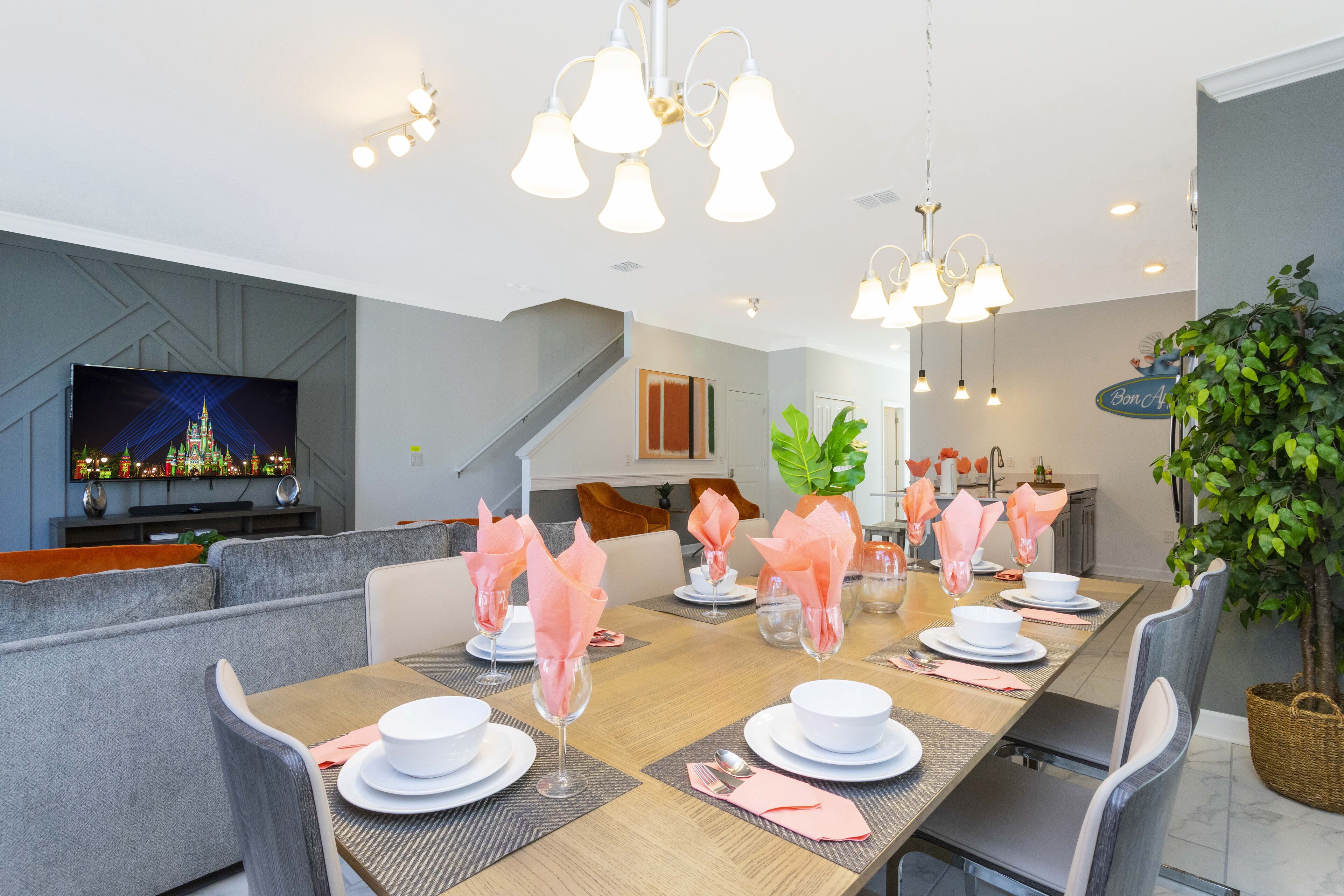 A dining area with a long table set for six, surrounded by grey upholstered chairs. A television is mounted on the wall to the left, and a staircase is visible in the background.