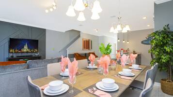 A dining area with a long table set for six, surrounded by grey upholstered chairs. A television is mounted on the wall to the left, and a staircase is visible in the background.