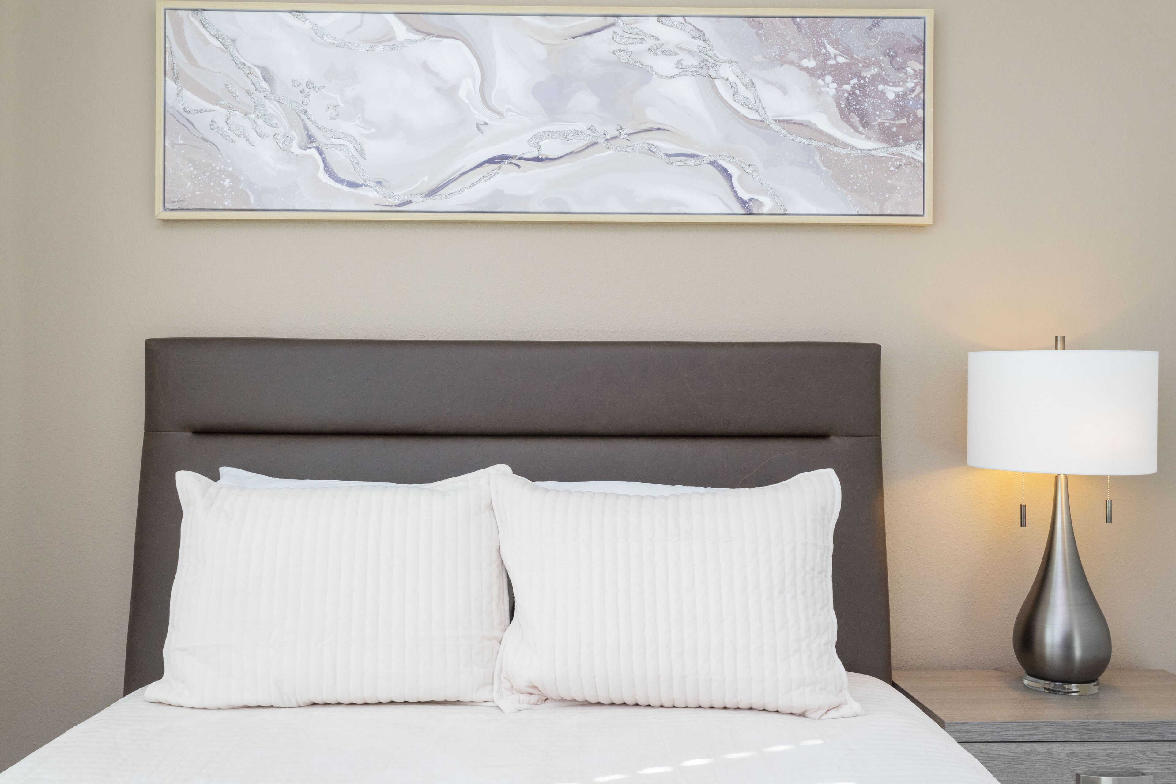 Un lit bien fait, avec deux oreillers et une tête de lit, une lampe de chevet et un tableau abstrait encadré accroché au mur.
