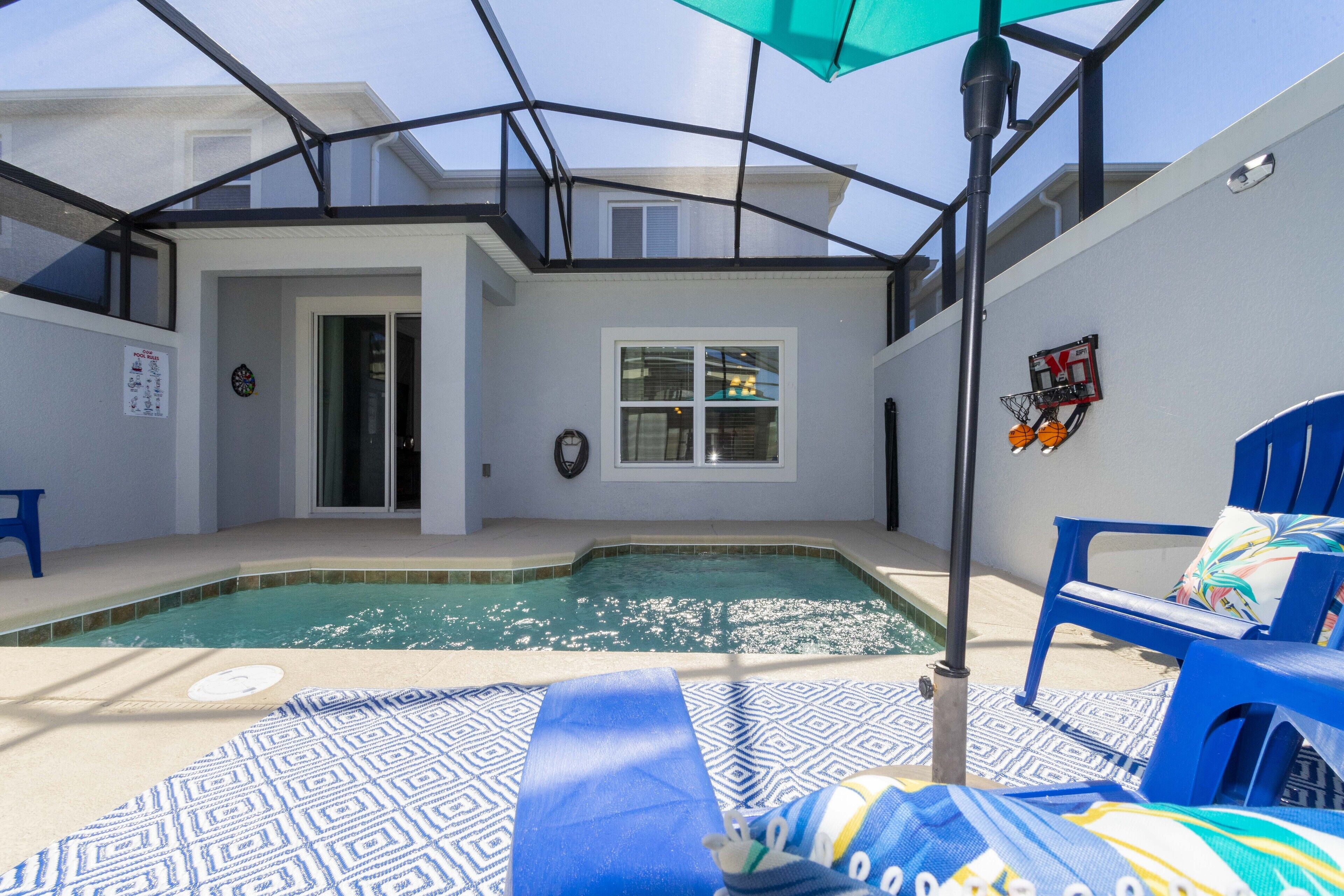 A pool area with a glass enclosure, a basketball hoop, and blue lounge chairs.